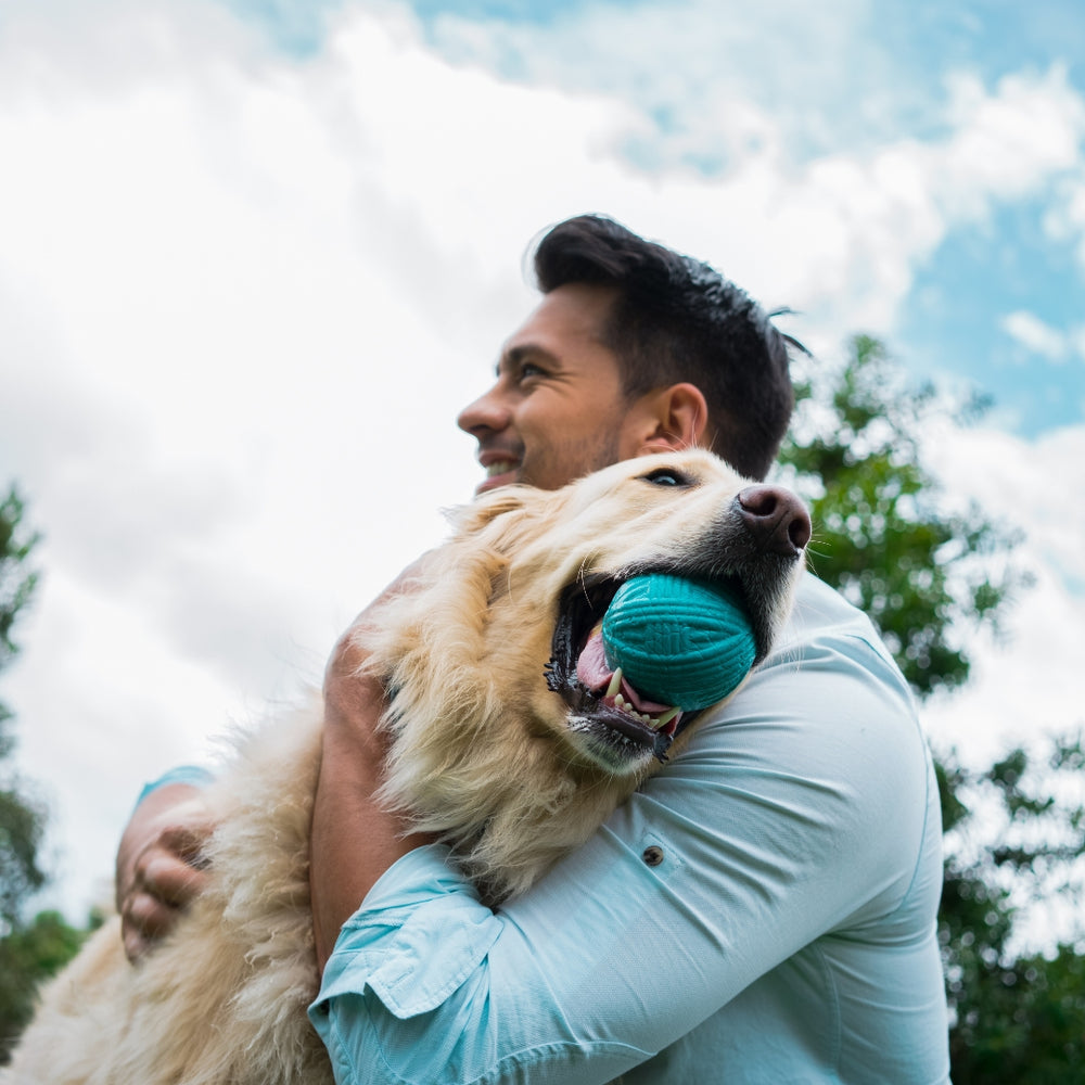 Man holding a dog with a blue ball in its mouth outdoors